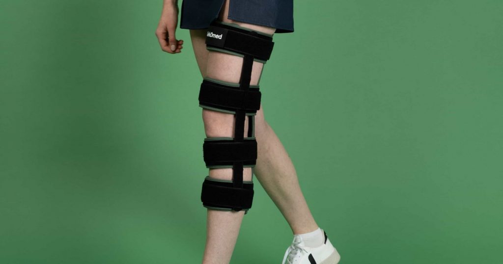 Femme portant une attelle d’immobilisation du genou Gekomed, debout sur fond vert, illustrant le maintien et l’ajustement du dispositif orthopédique.
